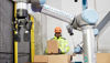 Worker in safety gear handling a box next to a robotic arm in a warehouse.