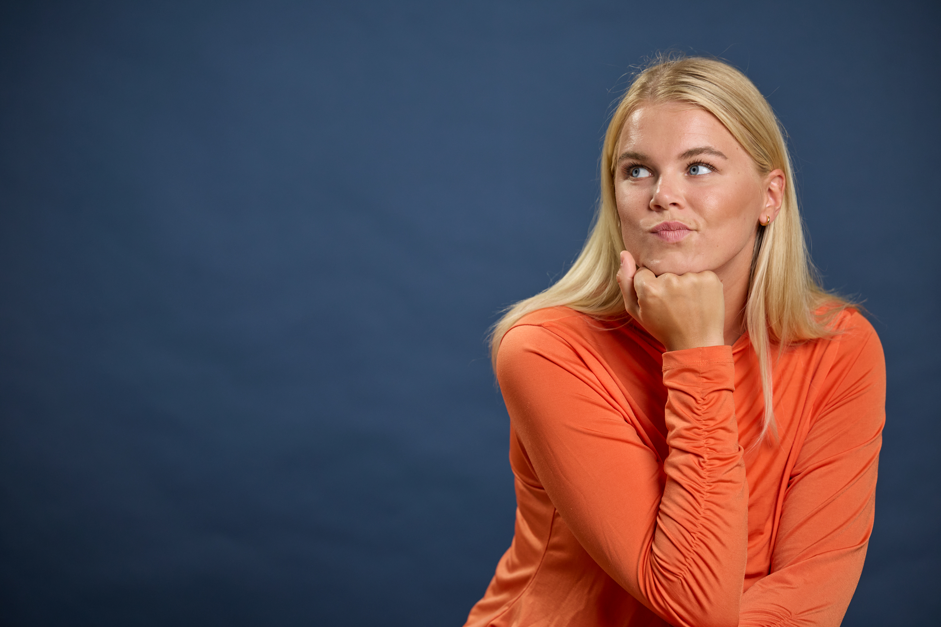 En blond kvinne i en oransje skjorte hviler haken på hånden og ser oppover tankefullt mot en mørkeblå bakgrunn.