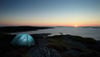 Ein blaues Zelt steht auf Felsen am Meer bei Sonnenuntergang