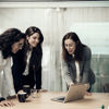 Three professional women in business attire focused on a laptop screen, engaged in a work discussion.