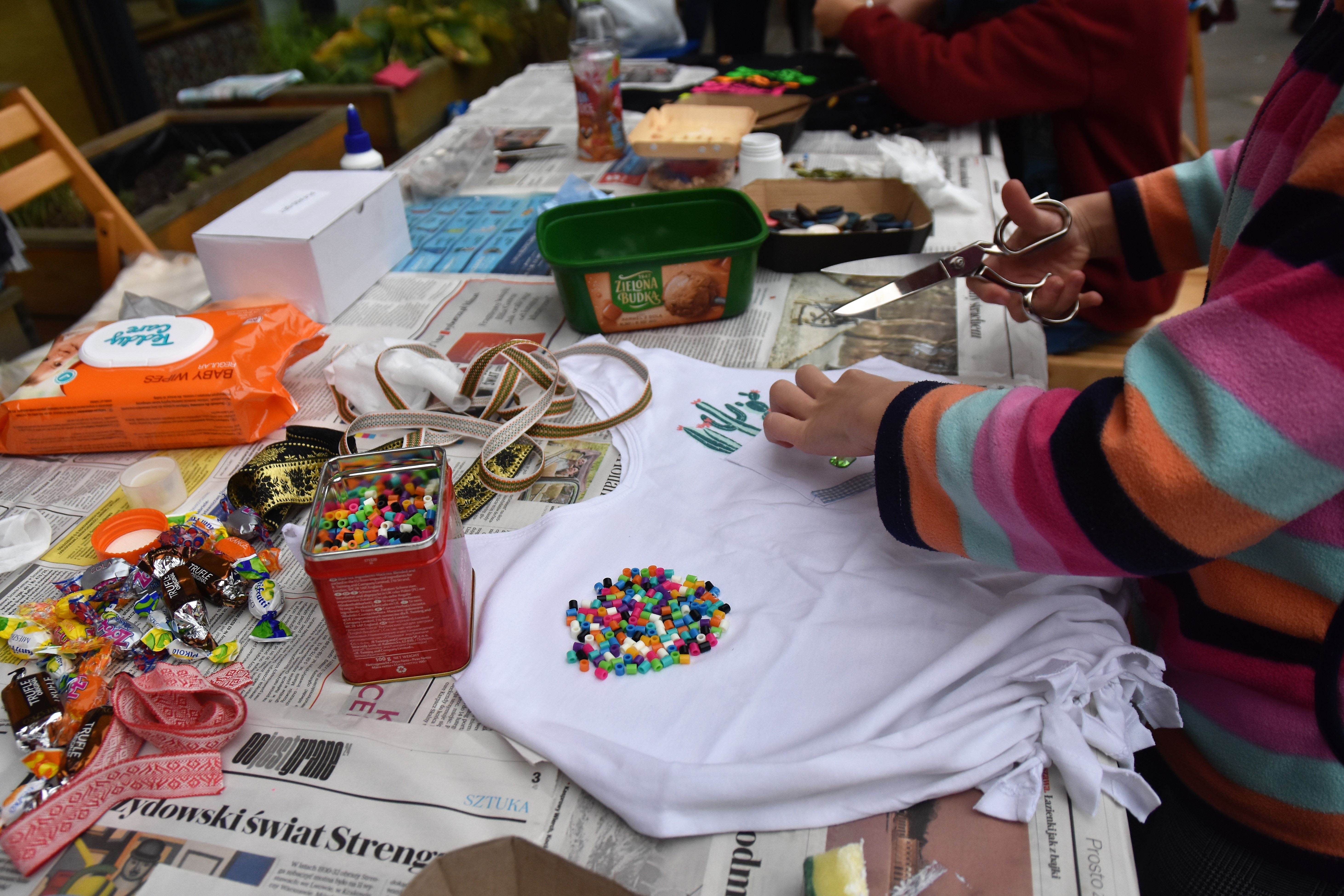 Zdjęcie przedstawia osobę, która przerabia i ozdabia pozornie niepotrzebną koszulkę, na stole znajduje się dużo ozdób