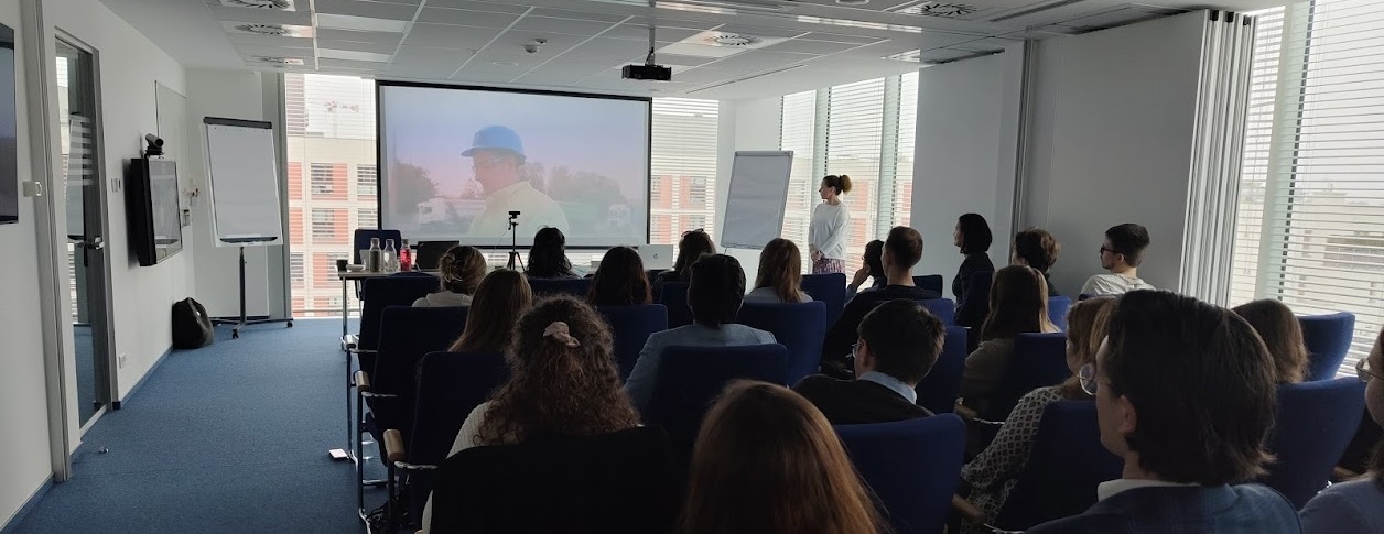 Na zdjęciu widać rzutnik, na którym wyświetlana jest prezentacja, osobę prezentującą oraz uczestników spotkania siedzących tyłem.