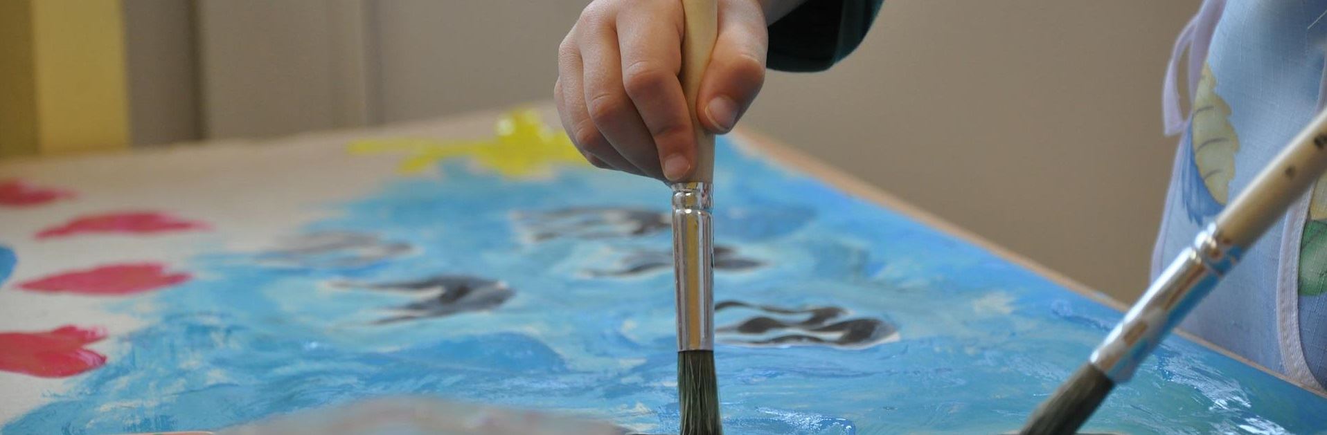 Niebieski obrazek oraz dziecięce ręce, które trzymają w pędzelki i malują