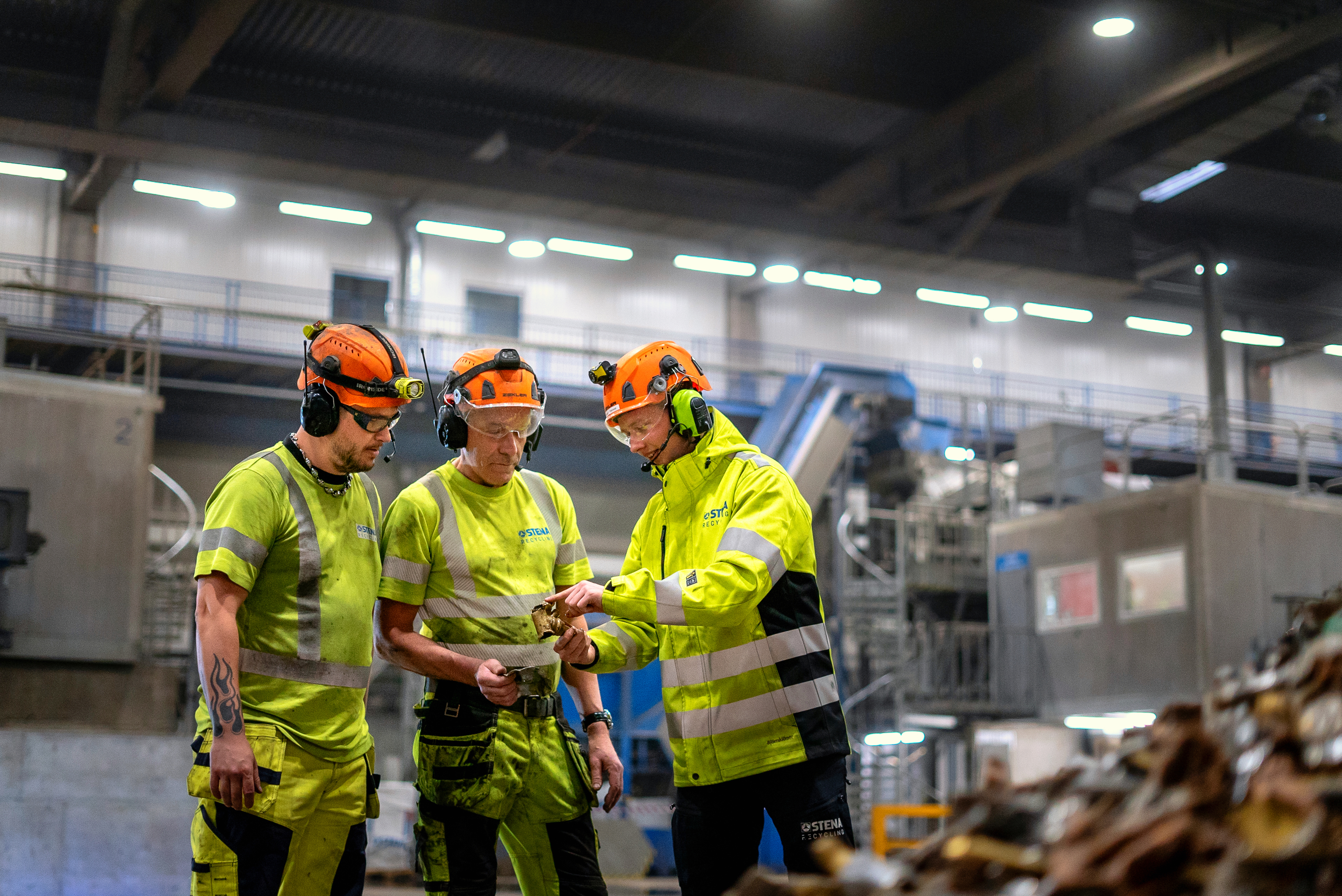 Drei männliche Stena Recycling-Mitarbeiter in Warnkleidung und Schutzausrüstung arbeiten zusammen im Stena Nordic Recycling Center.