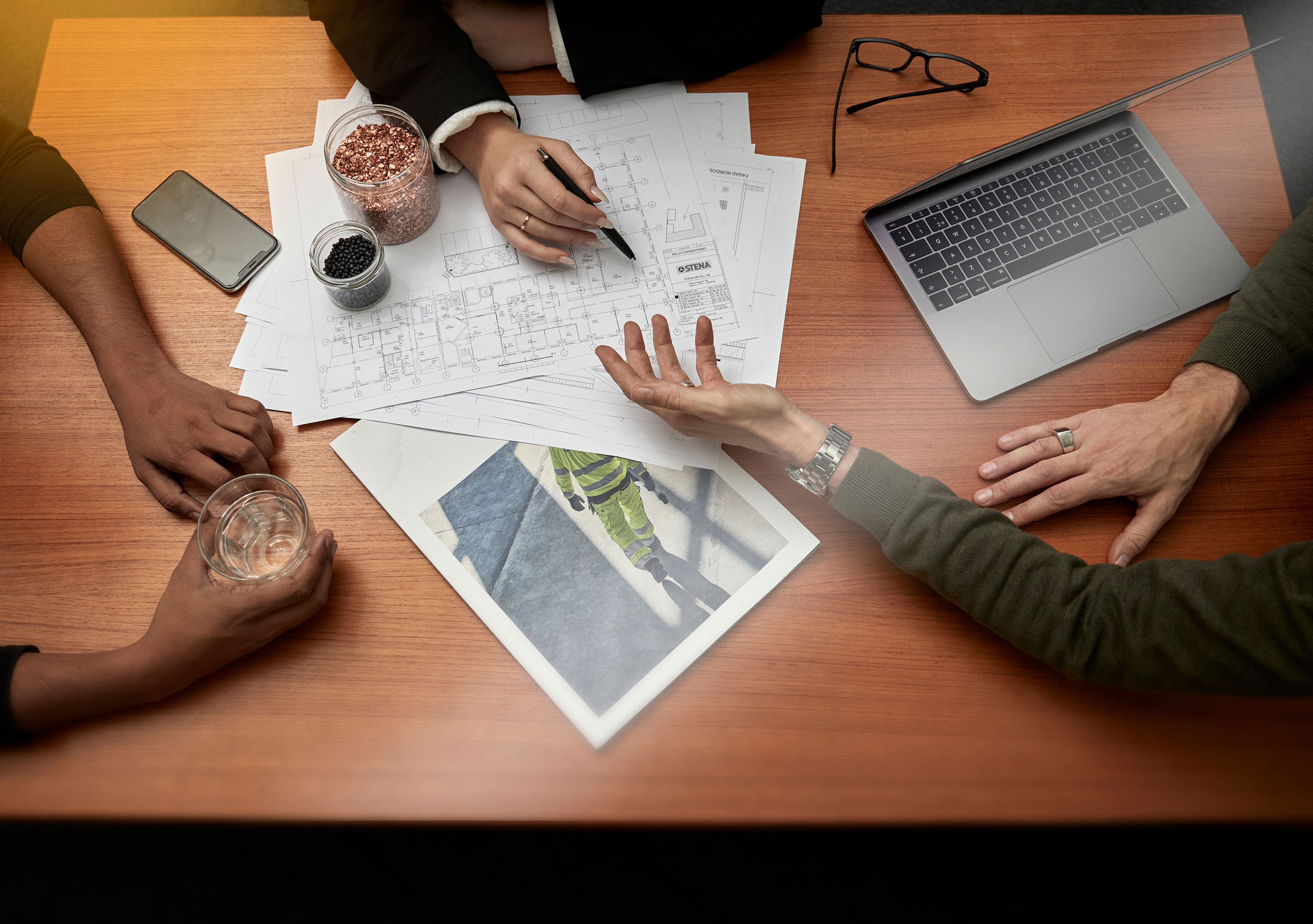 Händer på en träskrivbord med en bärbar dator, mobiltelefon, ritningar och två burkar av återvunnet material.