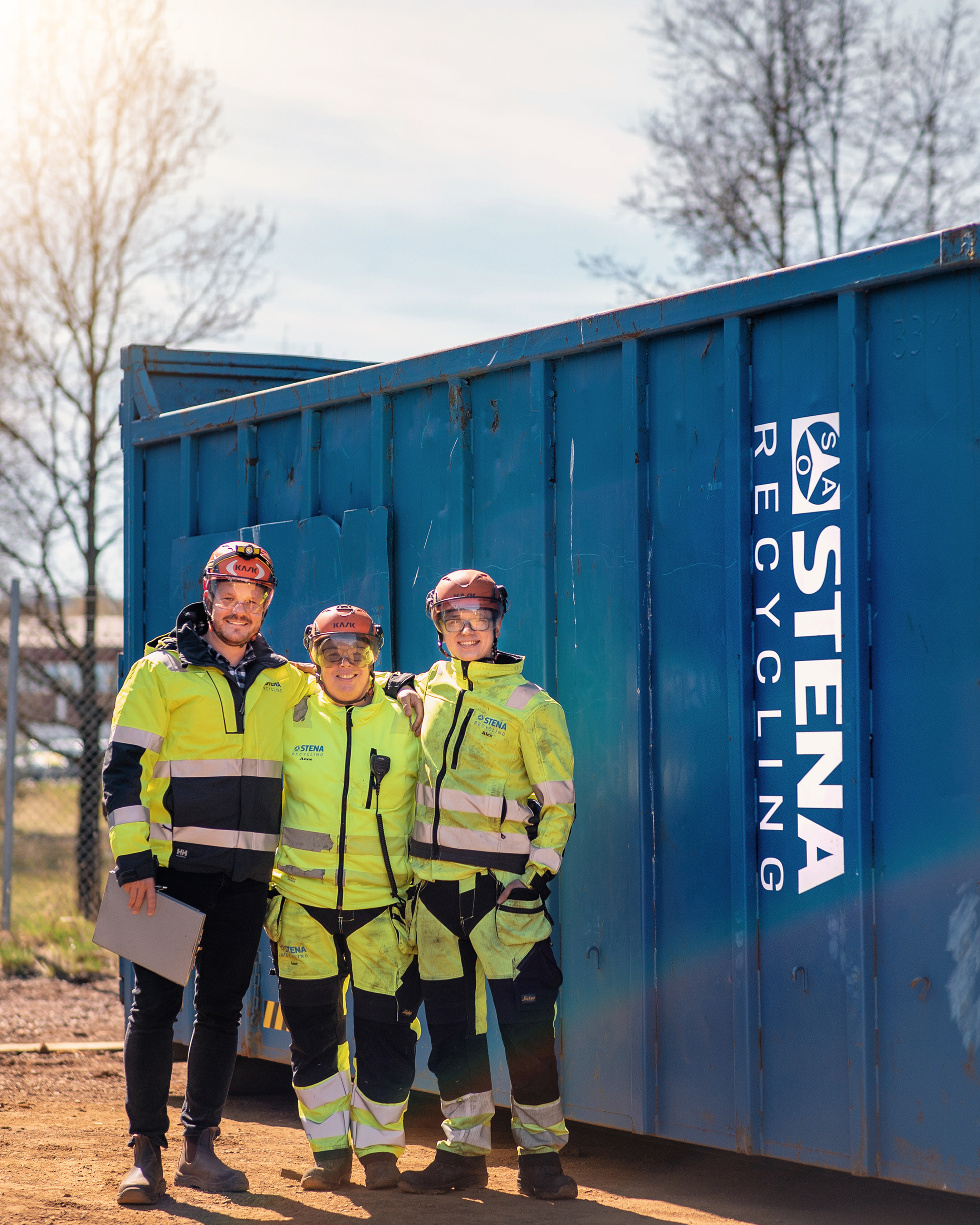 Three Stena Recycling experts dressed in reflective work-clothes, hard hats and goggles, standing in front of a blue Stena Recycling steel container, holding shoulders and smiling to the photographer