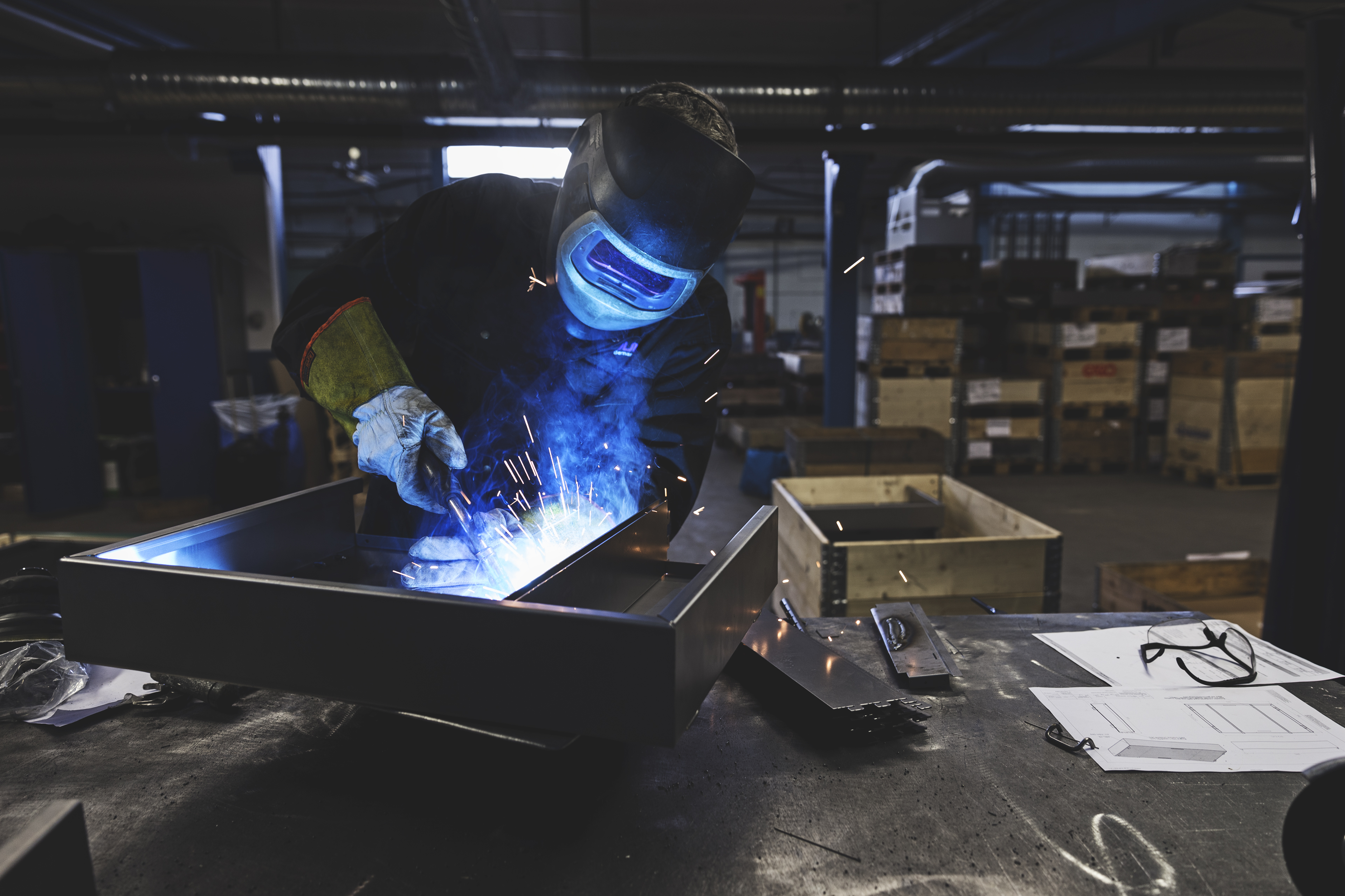 En arbeider i verneutstyr sveiser et metallstykke i et fabrikkmiljø.