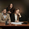 A female office worker looking over the shoulder of another female and a male colleague sitting at a desk, all watching a laptop screen.