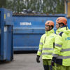 Two recycling experts dressed in hardhats and reflective Stena Recycling work clothes standing  beside a couple of large blue recycling containers in a recycling yard.