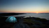 A blue tent standing on rocks by the ocean at sunset