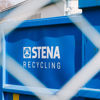 Blue metal container with the Stena Recycling logo, photographed through a chain-link fence at an industrial site.
