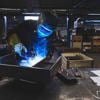 A worker in protective gear welds a piece of metal in a factory environment.