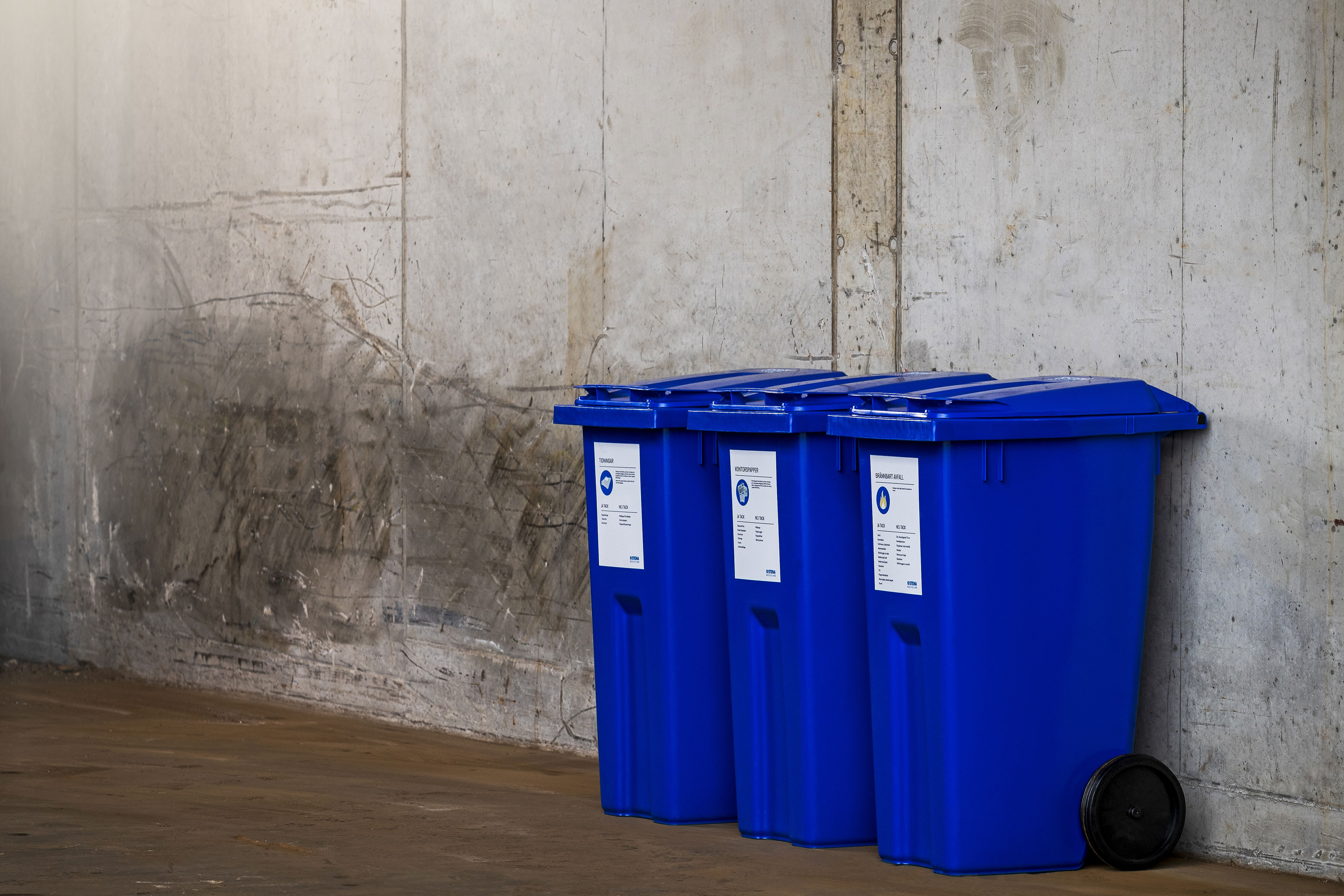 Three blue Stena Recycling waste collection containers placed in a row against a wall.
